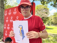 A participant wearing a red Special Olympics Venezuela shirt and hat stands outdoors in a park, holding a drawing of a colorful parrot. A Special Olympics Venezuela backdrop is displayed behind the participant, with trees, grass, and event attendees visible in the background.