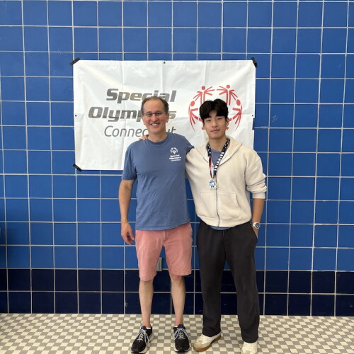 The photo shows an adult man standing next to a male high school student. They are in front of a blue wall with a Special Olympics Connecticut banner hanging on it. 