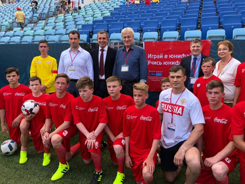A group of young people in a red kit kneeling in a line, one holding a football, with a line of adults standing behind them. Lines of empty stadium seats are in the background.