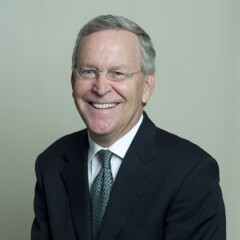 Jamie Aleman in front of a gray backdrop. he has on a dark suit and tie with a white button up shirt.