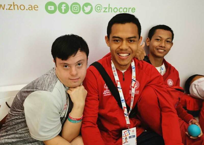 Four young men sitting in a group, two have on long sleep red Special Olympics Abu Dhabi 2019 pull overs. 