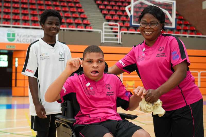 A smiling woman embraces her son, who is on a wheelchair, while her other son stands behind them, also smiling.