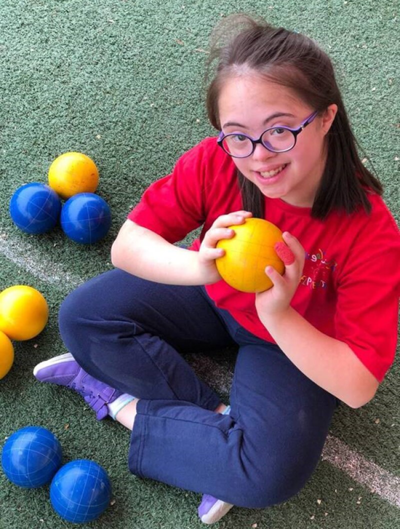 Sara Felemban sitting on a Bocce court holding a bocce ball looking up at the camera and smiling. 