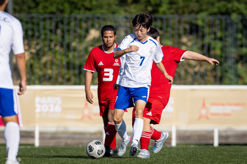 A football player wearing a white number 7 jersey controls the ball while two players in red attempt to challenge for possession on an outdoor pitch.