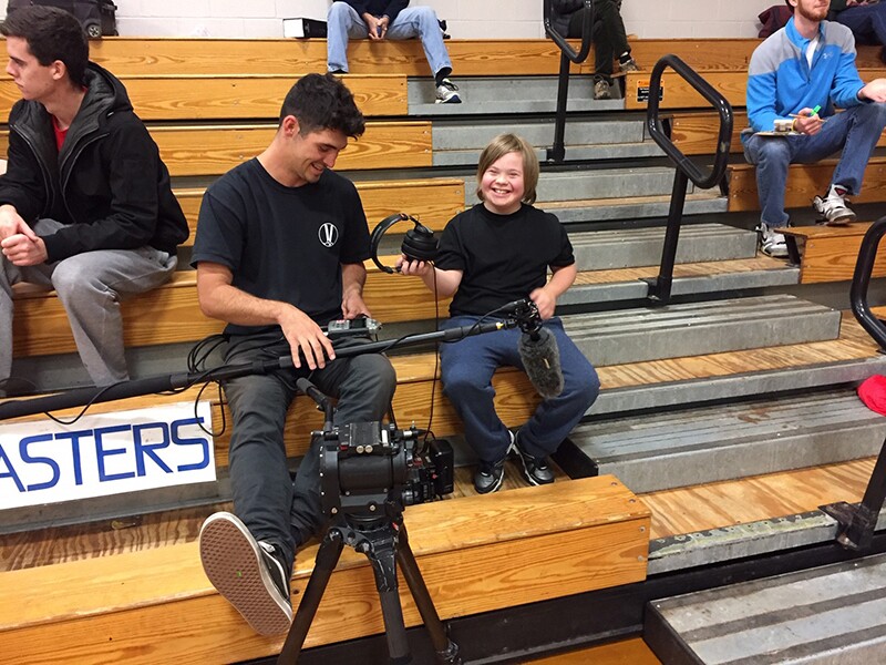 Dave, a videographer holing a boom mic and sitting next to Thorin on bleachers. Thorn is holding a head set and handing it to Dave.