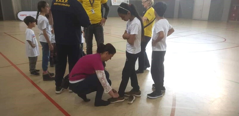 A woman is kneeling down to tie the trainer laces for a young girl who stands beside her. They are surrounded by a small crowd and are on an indoor court.