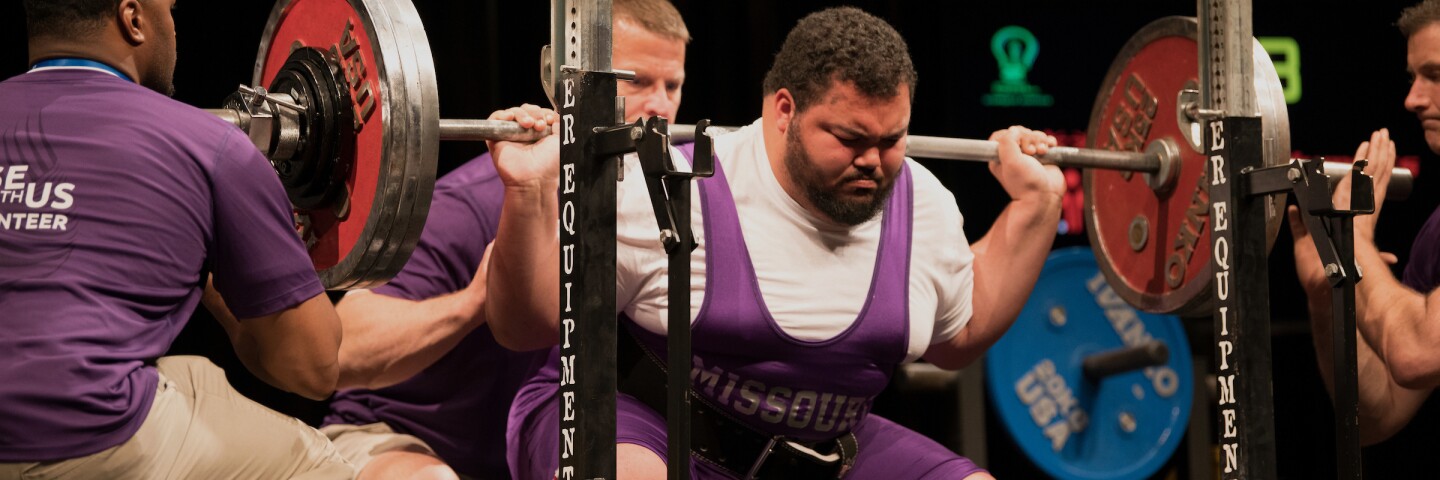 male lifter performs a squat at the rack while coaches and officials assist.