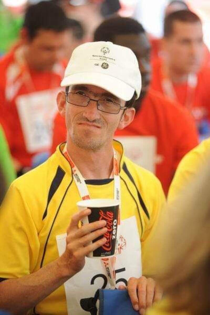 Man in yellow shirt and baseball cap drinks from a Coca-Cola branded paper cup.