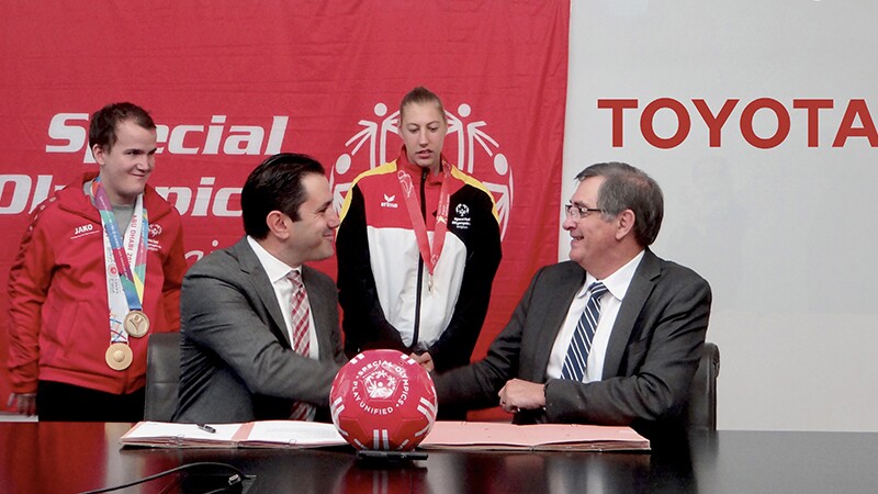 Two men sitting at a table shaking hands; the signing in the back ground (left) reads: Special Olympics; the signage in the background (right) reads: Toyota