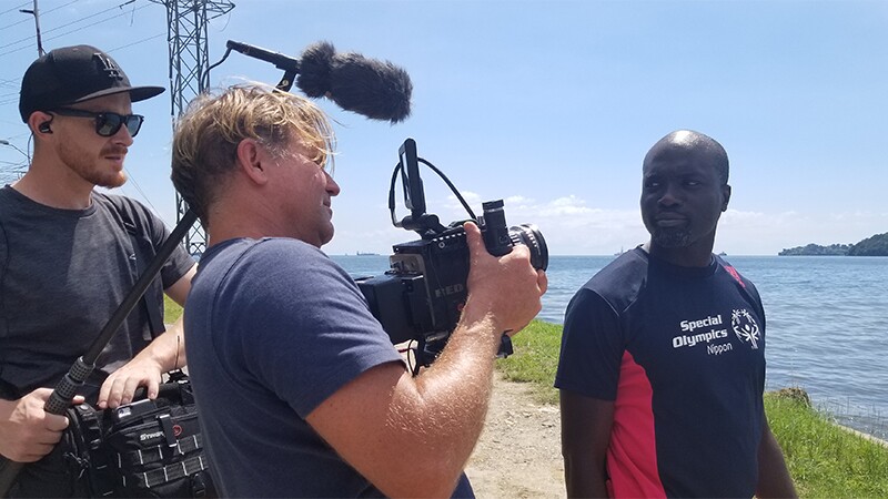 Kester being video recorded by a vidographer and boom mic operator. They are standing on a shoreline and water is in the background. Kester has on a Special Olympics Nippon black t-shirt.
