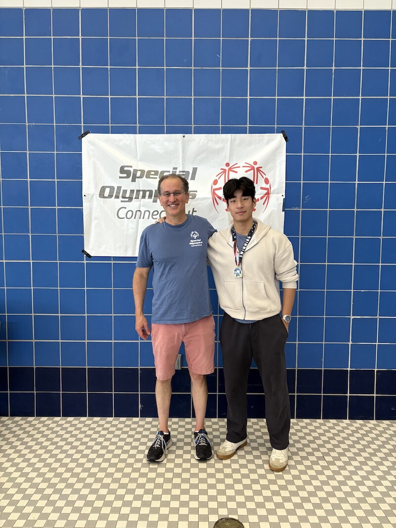 The photo shows an adult man standing next to a male high school student. They are in front of a blue wall with a Special Olympics Connecticut banner hanging on it. 