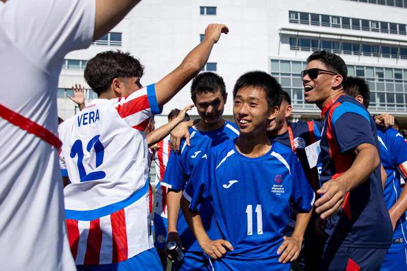 Football players in blue and white kits smile, congratulate one another, and exchange high-fives with coaches on the field after a match.
