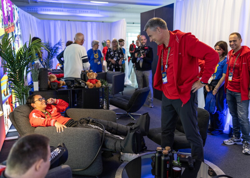 In a busy room full of people, a man in a recliner chair smiles up at a man standing near him.  