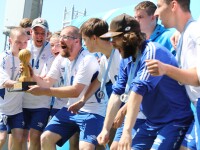 A group of men in blue shorts and white t-shirts stand together around one man who is holding a trophy. They have their mouths open as if shouting or cheering.  
