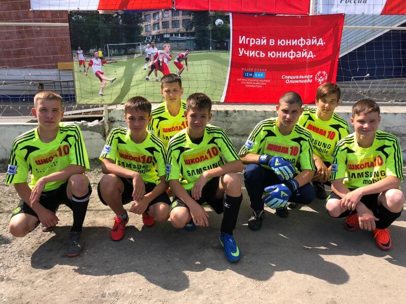 Seven boys in luminous yellow football jerseys, black shorts and football boots kneel in front of a banner branded by Special Olympics and Stavros Niarchos Foundation.