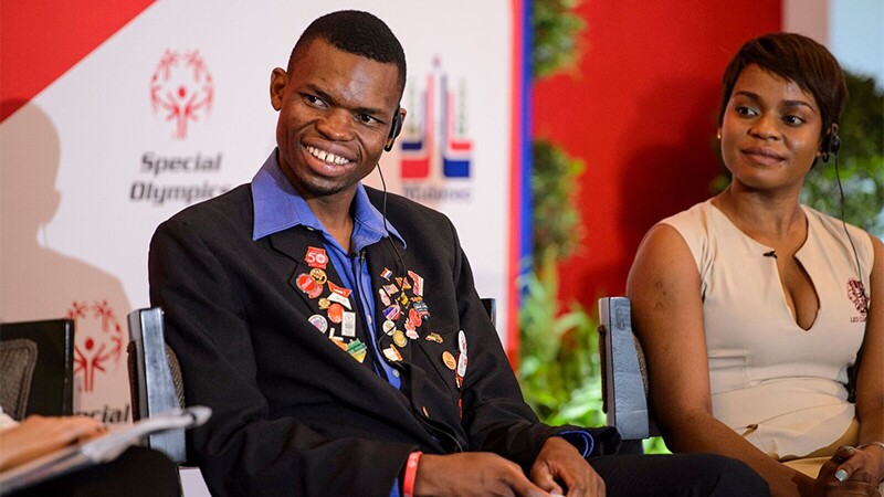 Nyasha Derere from Zimbabwe speaks on stage as a panelist during the Global Athlete Congress in Santo Domingo, Dominican Republic. He has on a blue collard shirt with a black jacket that is adorned with Special Olympics and iInclusion pins. A representative is sitting to his left.