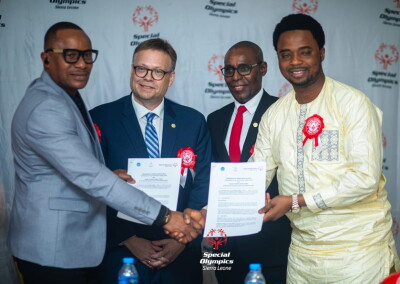 Officials exchange signed documents and shake hands during a Special Olympics partnership signing event in Sierra Leone, with branded backdrop visible.