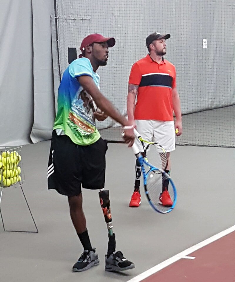 Dionte Fosters plays tennis with his new prosthetic leg; he just took a swing at a ball. Jeff Bourns is in the background watching while holding his racket and ball.