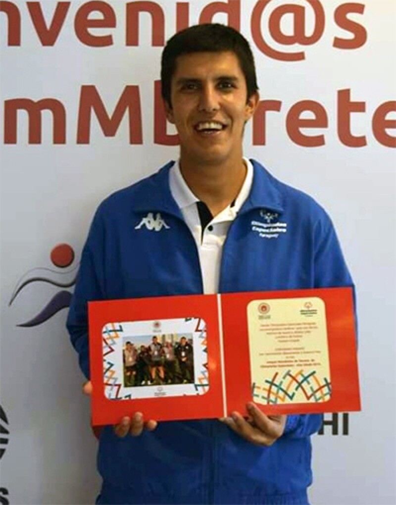 Hassam stands in front of a step and repeat that says “Secretaria Nacional de Deportes.” He is holding a microphone.