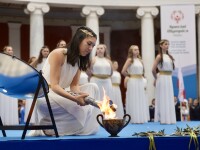 A woman in a white dress and golden head piece kneels down holding a lit torch.