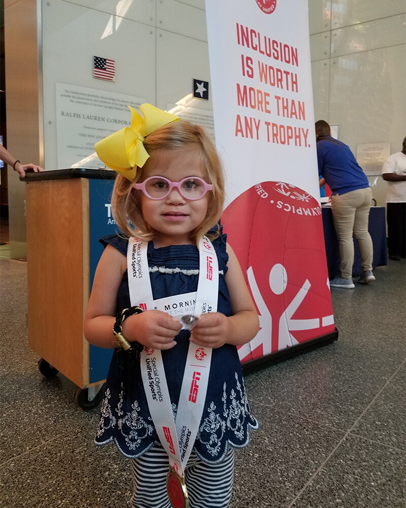 A young girl wearing pink glasses and a blue dress with white and blue horizontal striped leggings tries out a Special Olympics medal at the museum’s hands-on section. She is standing in front of a Special Olympics sign banner that reads, Inclusion is worth more than a trophy.