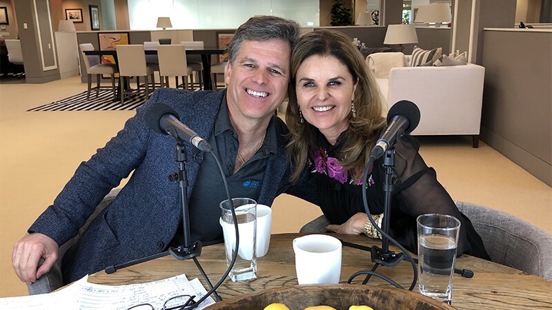 Tim & Maria Shriver sitting next to one another at a table with two mics in front of them.