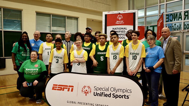 Group of students and faculty standing in two rows in the gymnasium next to Unified Champion School signage.