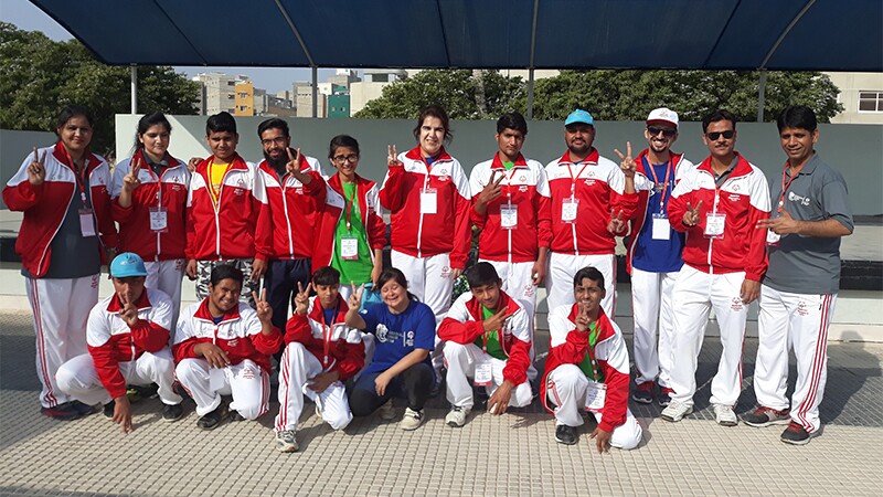 Jasmine poses with athletes from Special Olympics Pakistan. They are wearing matching tracksuits, and many of them are doing peace signs with their hands.