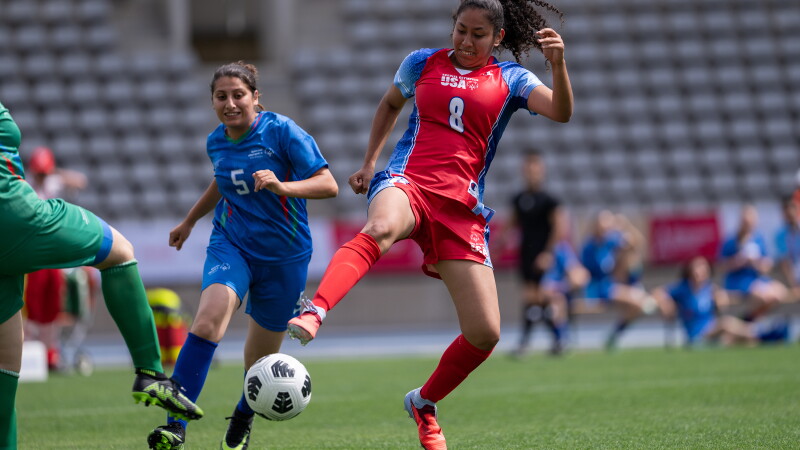 A Team USA player in a red jersey controls the ball during a football match as a defender in blue closes in, with the goalkeeper and stadium seating visible in the background.