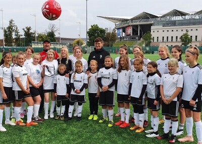 A group of women and girls in football kit stand together on a green pitch while a red football bounces in the air above them.