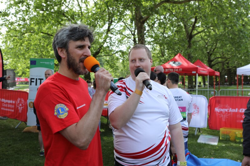 Two men with microphones standing in front of trees and pull-up banners in a park.