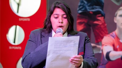 Young woman reading from a script and speaking on stage. 