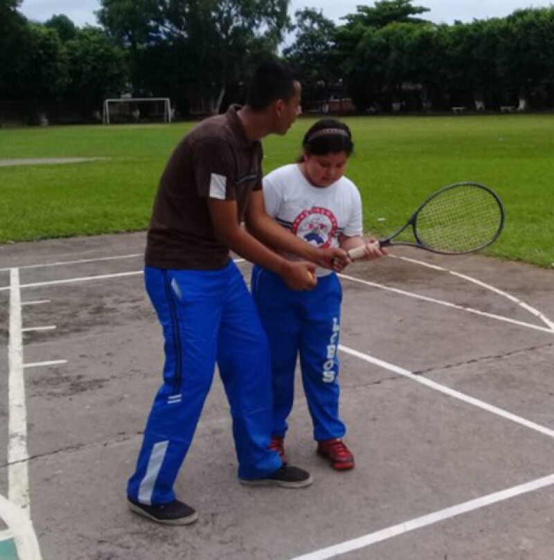Instructor is teaching a young girl on the court; the instructor is teaching the young girl how to hold a tennis racket.