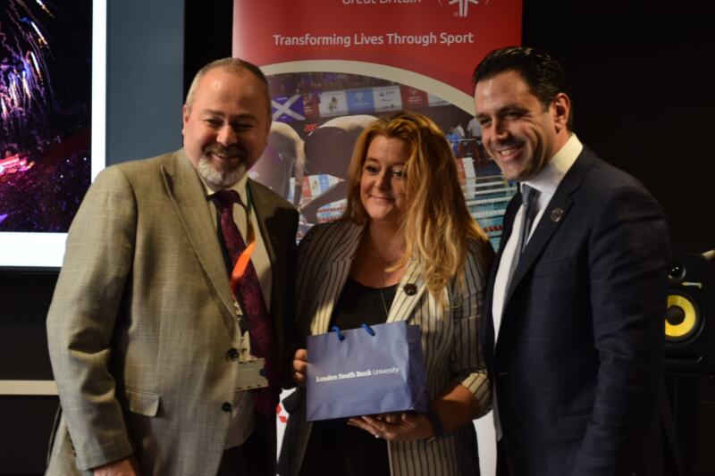 A woman stands between two men holding a present and smiling for the camera with Special Olympics branding in the background. 