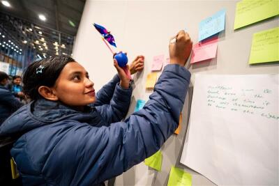 A young woman wearing a winter jacket writes notes on colorful sticky notes posted on a large board in a busy indoor event space.