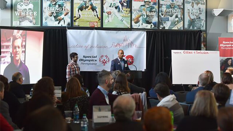 Two presenters on stage speaking to an audience with America football and Special Olympics materials displayed on the stage.