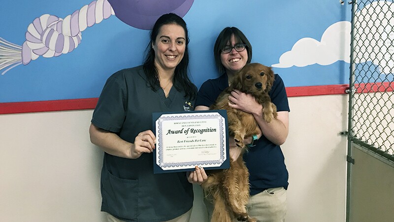 Katy Sanchez (R) is holding a small dog and standing next to a colleague that is presenting Katy with an Award of Recognition.