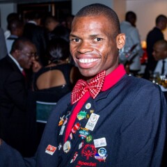 Nyasha Derera sitting at a formal event wearing a dinner jacket, red bow tie, and inspirational pins on his lapel. He's smiling and giving a thumbs up.