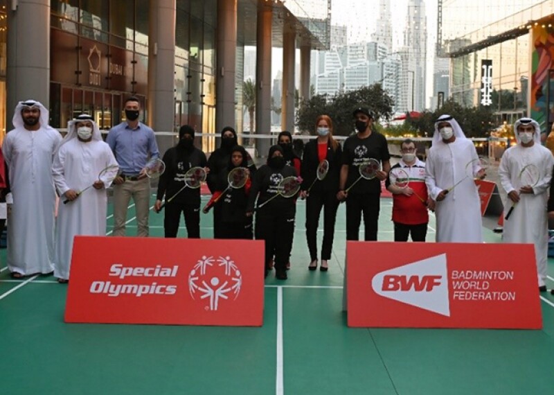 A group of people standing behind A-Board signage and a badminton court with large buildings in the backdrop.