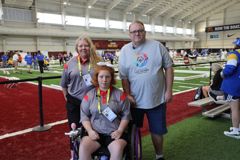 Emileigh Marsh sits in a wheelchair on an indoor bocce competition field at the 2026 Special Olympics USA Games, flanked by Kelly Marsh and Bobby Marsh. All three pose together near the bocce courts, with athletes, volunteers and competition areas visible in the background.