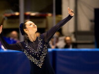 A figure skater is pictured performing a routine, her arms raised artistically. Blurred behind her are spectators. 