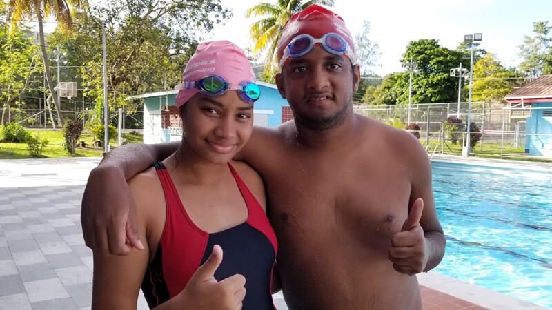 Shania and Stefan (Open Water Swimming) just got out of the pool and are posing for a photo together; both are wet, in their swimming suits, caps, and goggles; both are smiling and giving a thumbs up. 