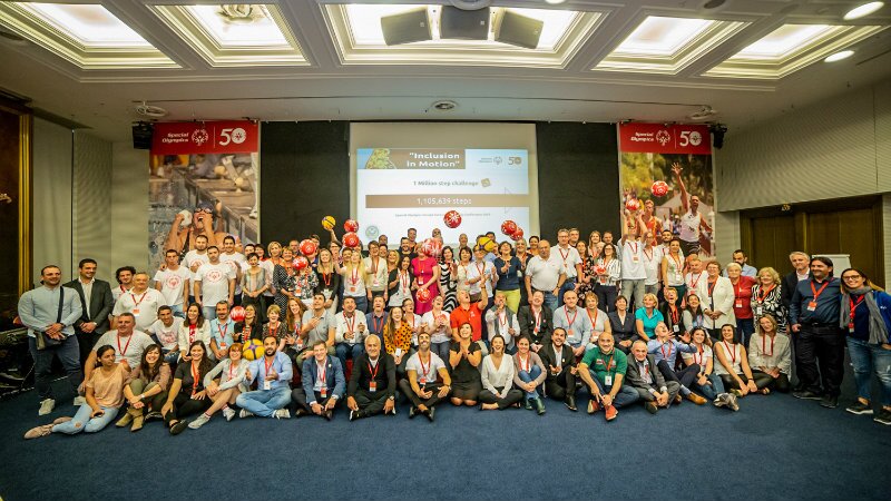 Around 100 conference participants gathered in front of a large screen and Special Olympics branding.