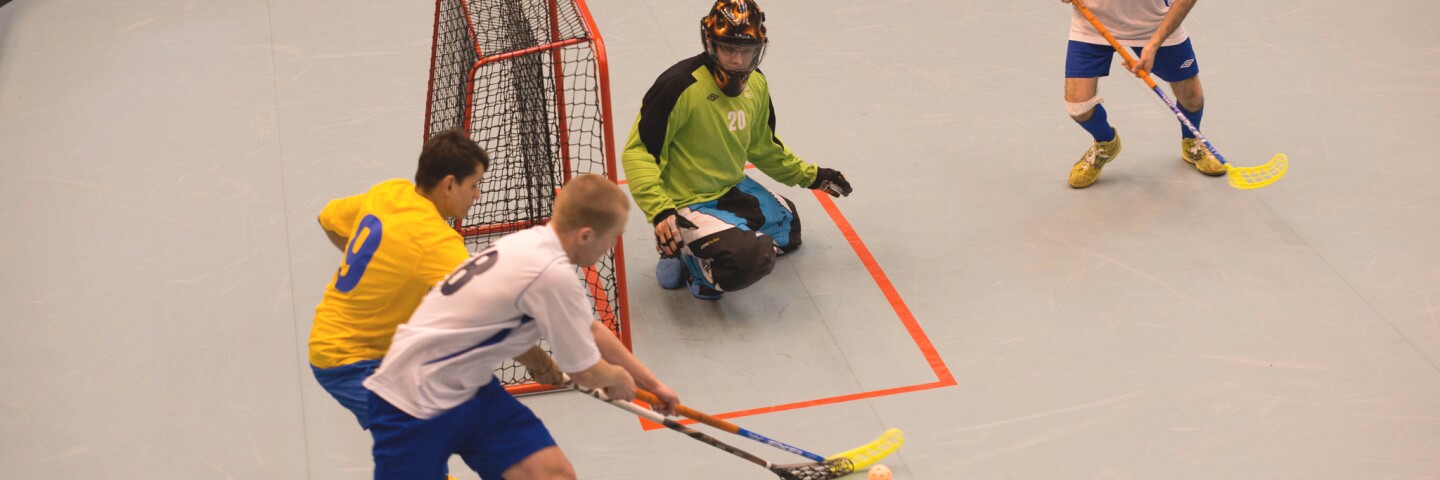 Athlete attempting to make a goal while his opponent blocks; the goalie and another player watch.