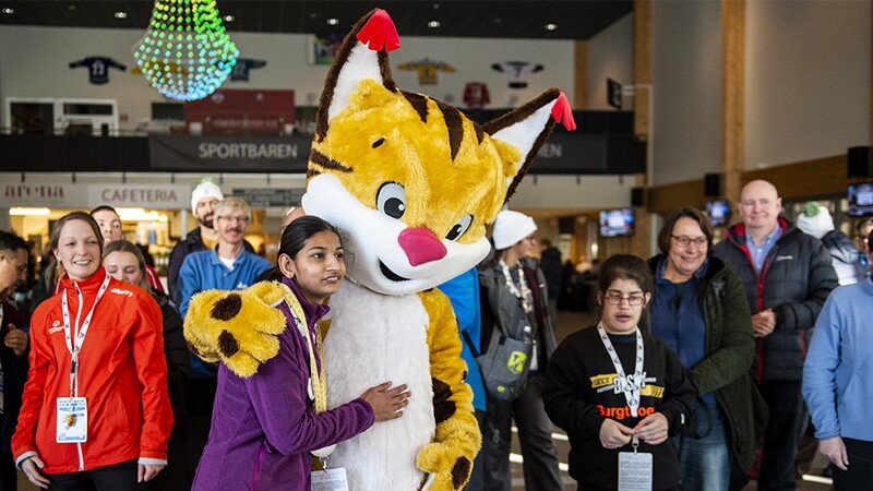 Speed Skater, Rachna, standing next to the cat mascot from the Sweden Invitational Games posing for a photo together while people stand around them and look on.