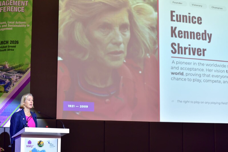 A woman stands behind a podium while she speaks during a presentation. A large screen is behind her showing a PowerPoint presentation.