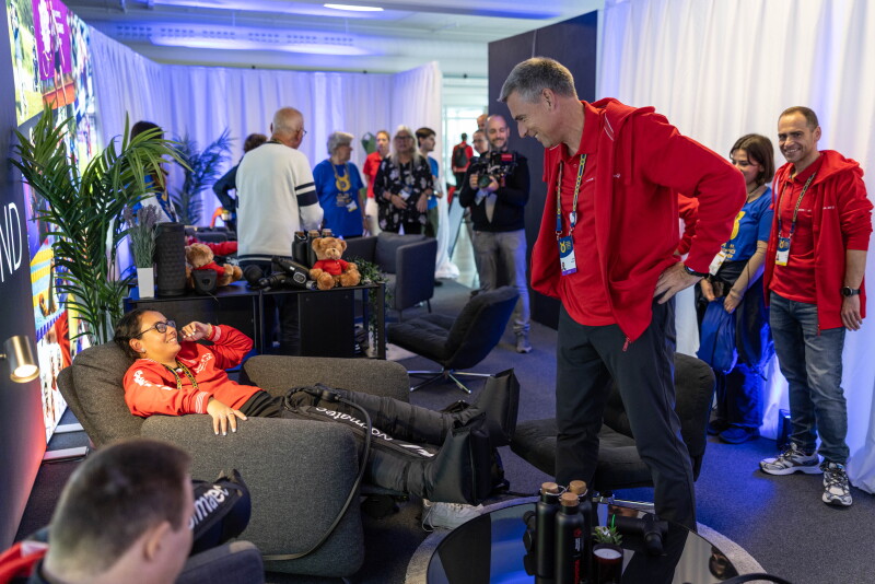In a busy room full of people, a man in a recliner chair smiles up at a man standing near him.  