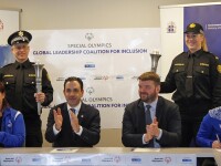 One woman (clapping) and three men (two clapping and one signing a document) sit at a table which is branded with Special Olympics and SNF logos.