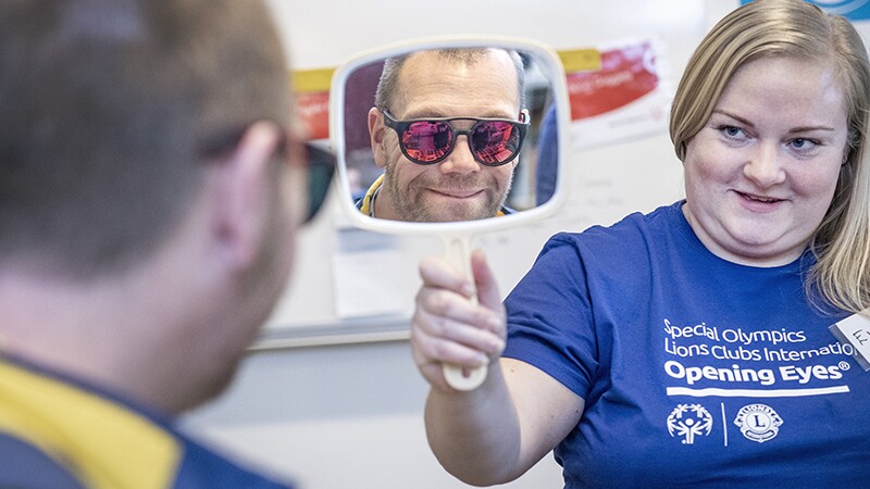 Athlete trying on ski glasses as a volunteer shows him his reflection in a handheld mirror.