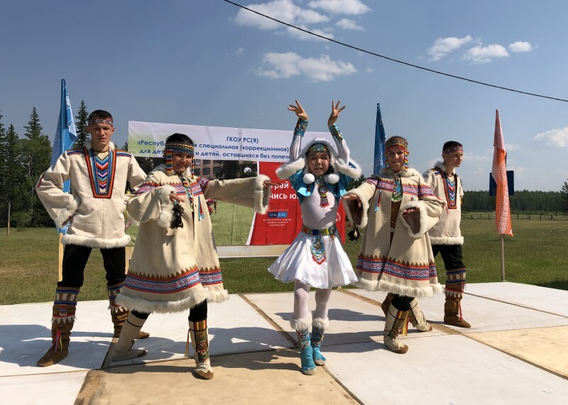 Five young people in traditional costumes pose on a stage with grass and a blue sky in the background.